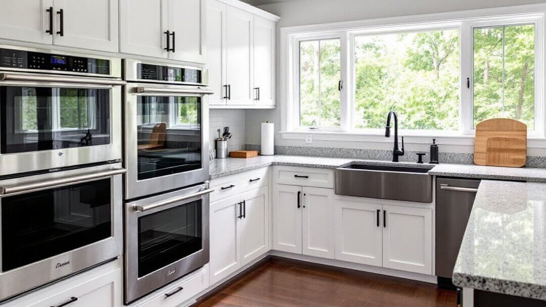 Modern kitchen with a bright interior featuring double stainless steel convection and traditional ovens built into white cabinetry. Large windows provide natural light, and the space includes a farmhouse sink and granite countertops.