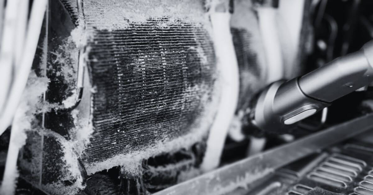 Close-up of dusty refrigerator coils being cleaned with a vacuum cleaner attachment, showing the buildup of dirt and debris being removed.